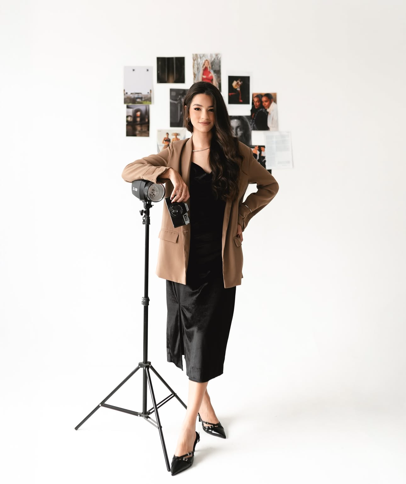 Woman in black dress and tan blazer poses beside a camera tripod with a mood board in a bright studio.