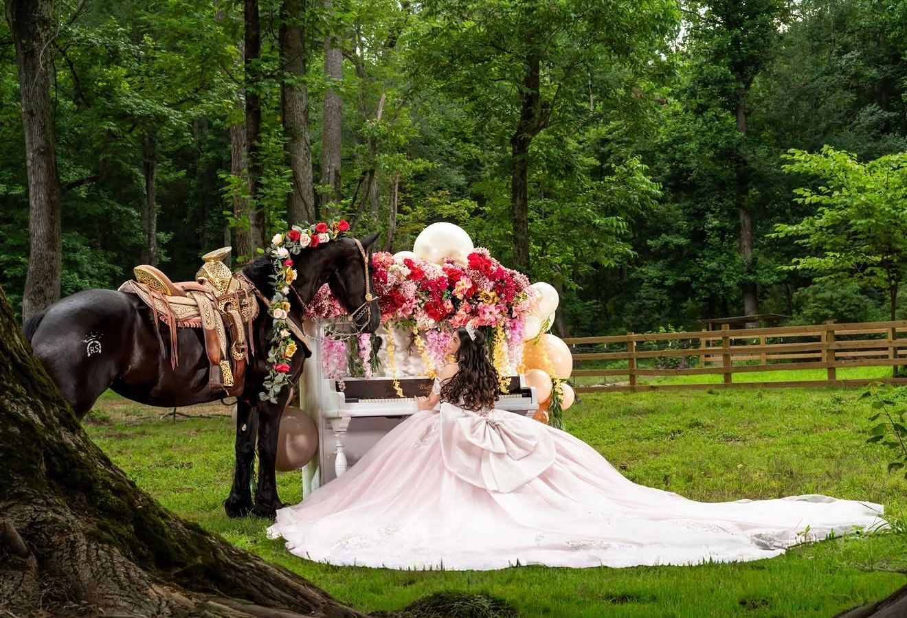 A bride in a pink gown poses with a floral-adorned horse in a lush green forest setting.