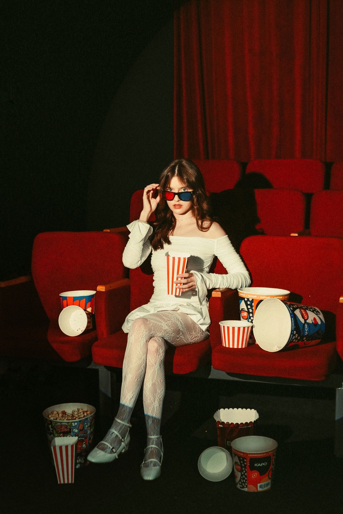 Young woman in 3D glasses sitting in red cinema seats surrounded by popcorn buckets and snacks in a movie theater.