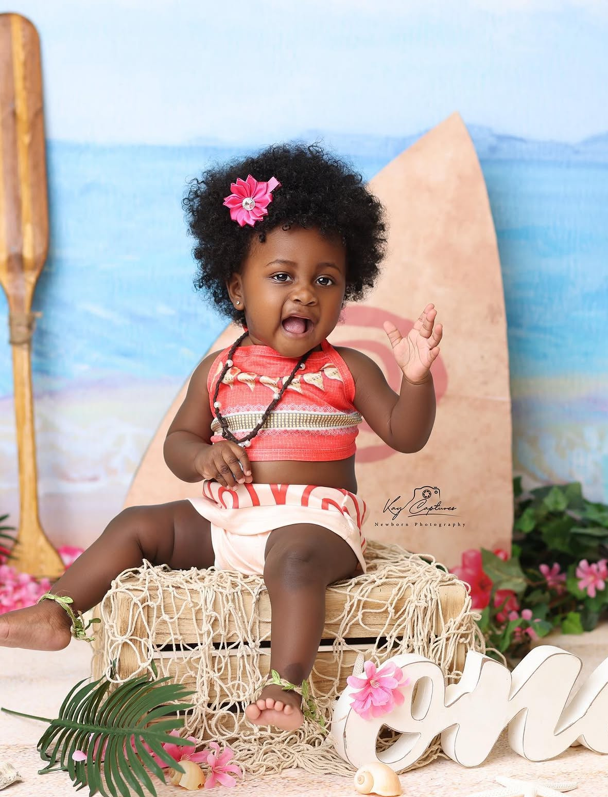 Adorable baby girl in coral Moana costume sits on wicker box at tropical themed first birthday photo shoot.