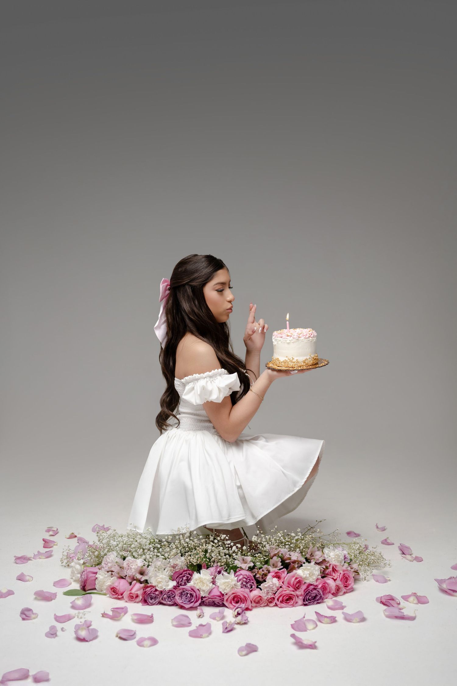 Young girl in white dress holding birthday cake with candle, sitting among pink and white roses on gray background.
