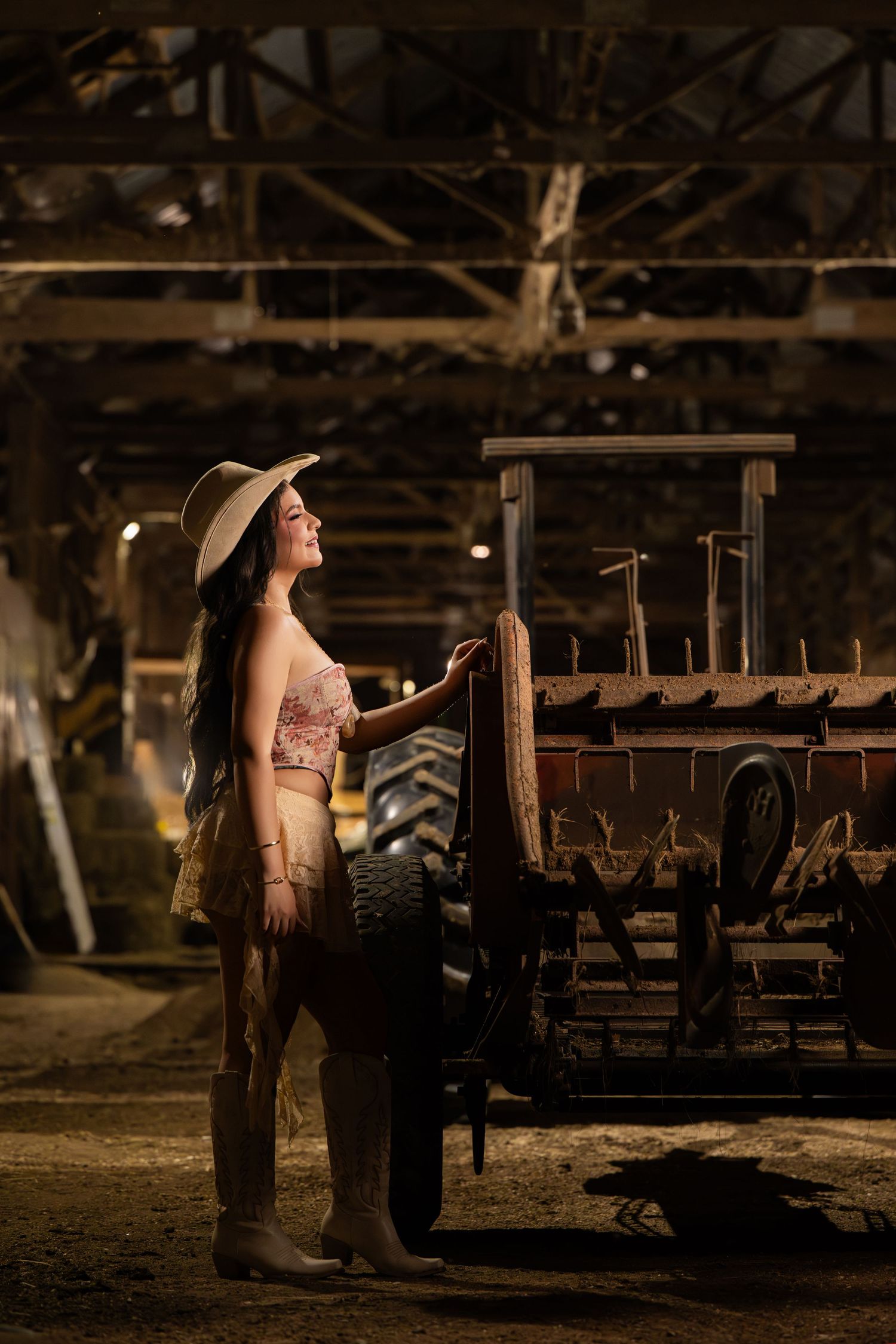 Woman in cowboy hat and western outfit stands beside rustic farm wagon inside a dimly lit barn.