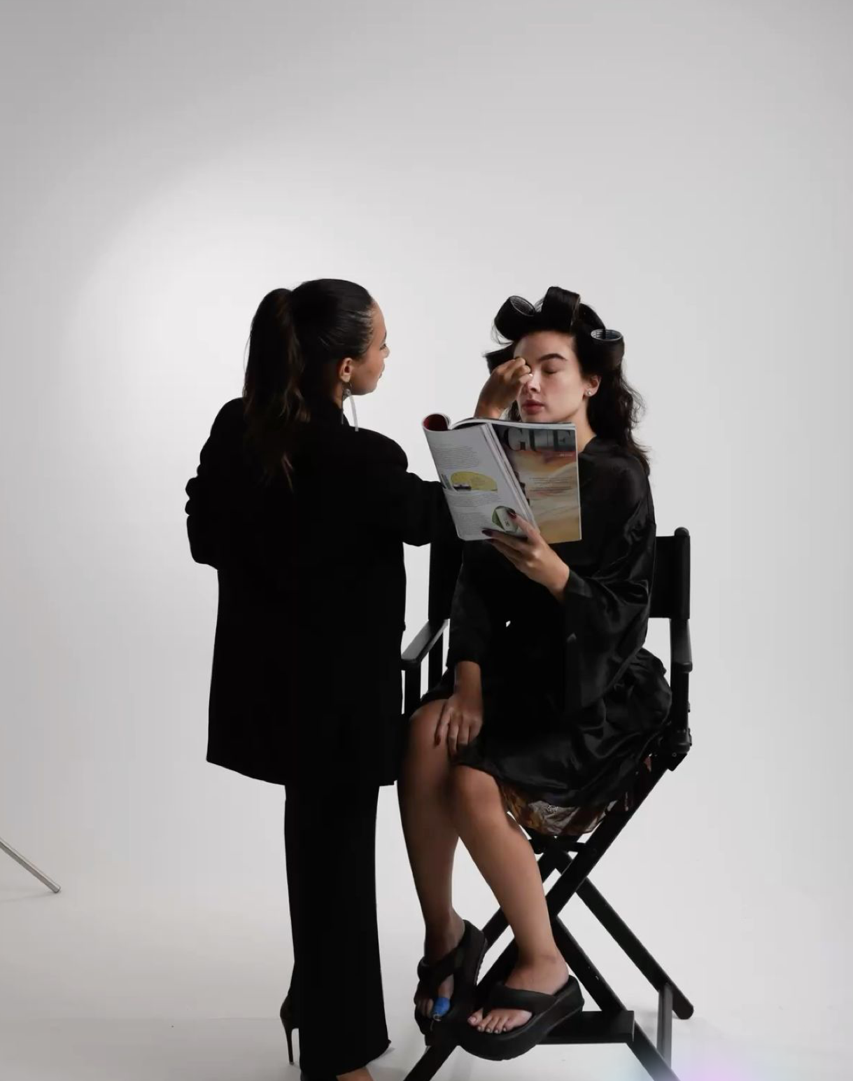 Makeup artist applying makeup to a seated model with hair rollers reading a magazine in a studio.