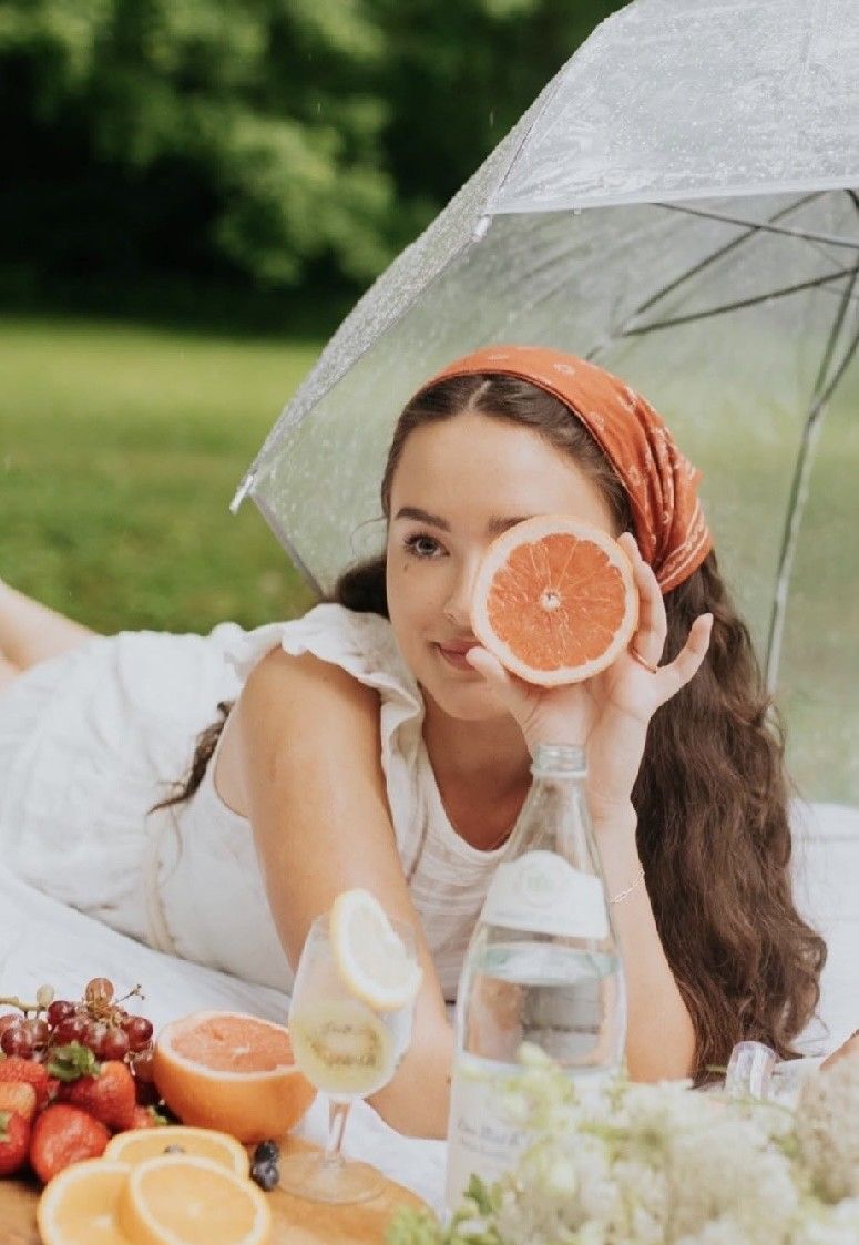 Woman holding a grapefruit half at a picnic with fruits, water, and a clear umbrella outdoors.