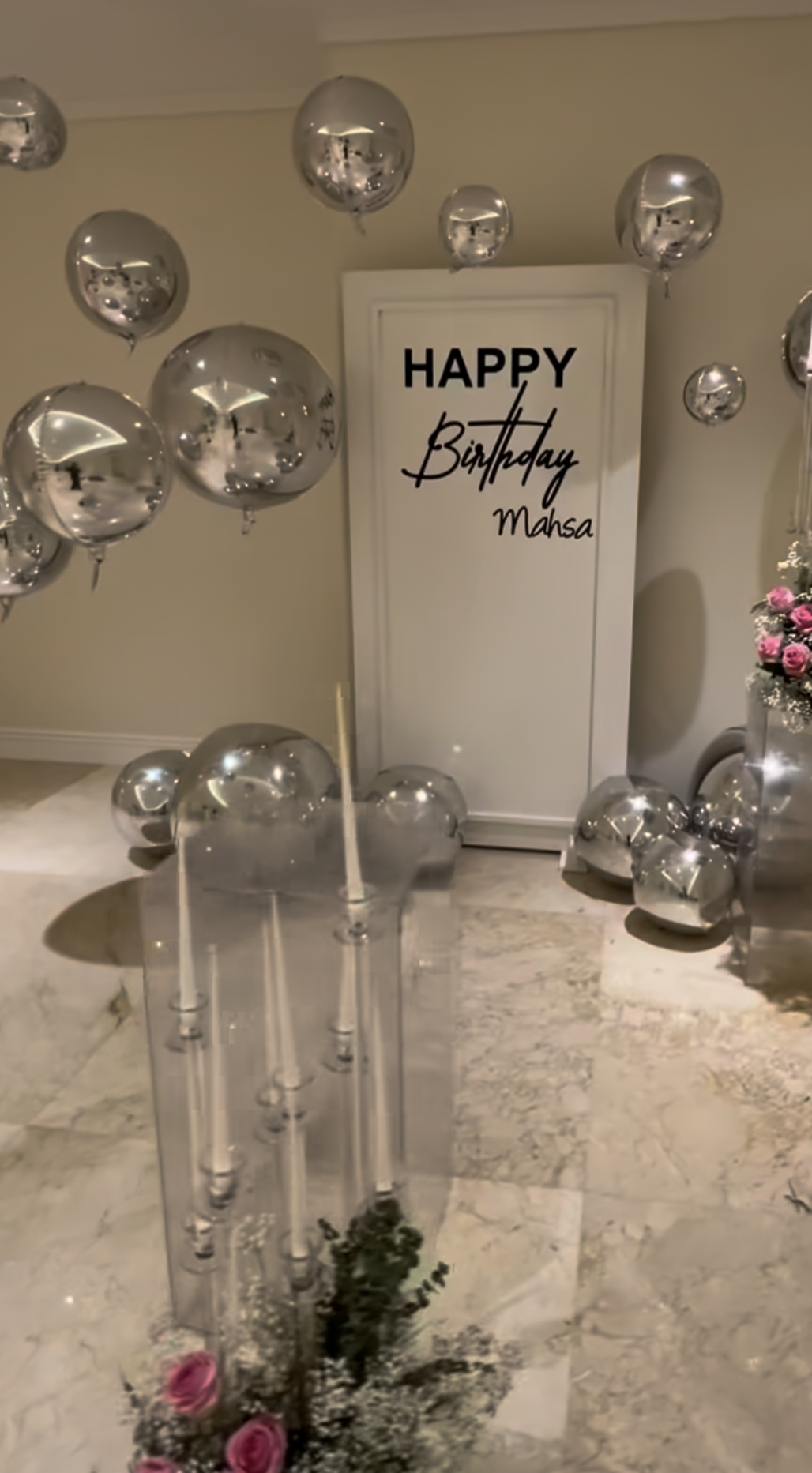 Silver balloon birthday decoration with Happy Birthday Mahsa written on white door, candelabra and pink roses.