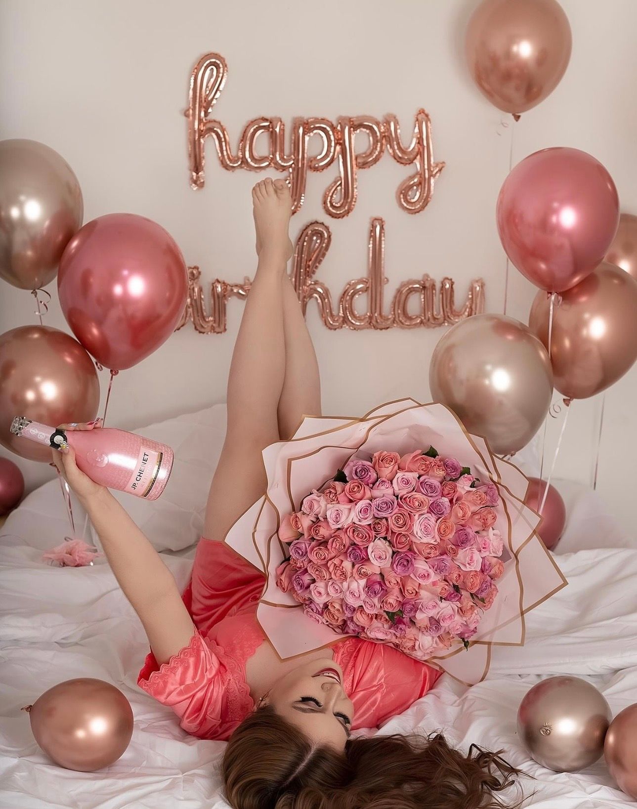 Woman lying on bed with pink roses bouquet, champagne bottle, rose gold balloons, and happy birthday foil letters.