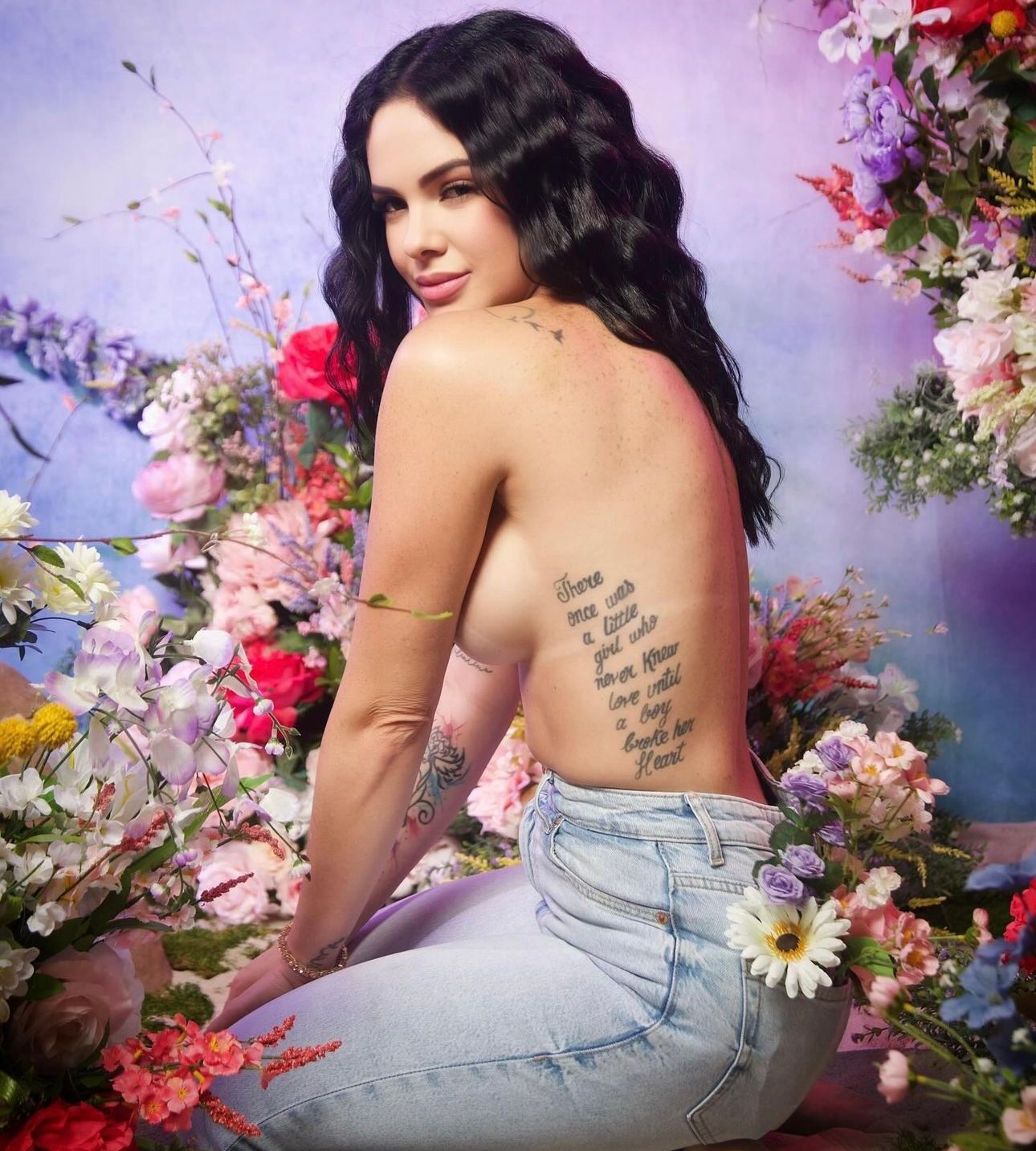 Woman with dark wavy hair and back tattoo poses topless in jeans among colorful flowers.
