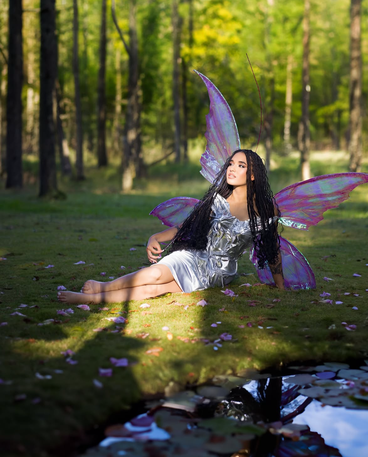 A woman in a silver dress with purple fairy wings sits gracefully on mossy ground in a sunlit forest.