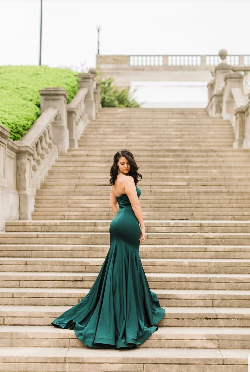 Woman in elegant emerald green mermaid gown poses on grand stone staircase, looking back over her shoulder.
