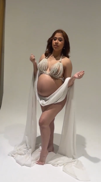 A pregnant woman poses in white flowing fabric and beaded lingerie for a maternity photoshoot against a light backdrop.