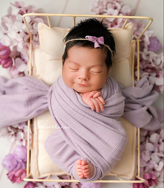 Sleeping newborn wrapped in purple swaddle blanket lying in gold frame with floral decorations.