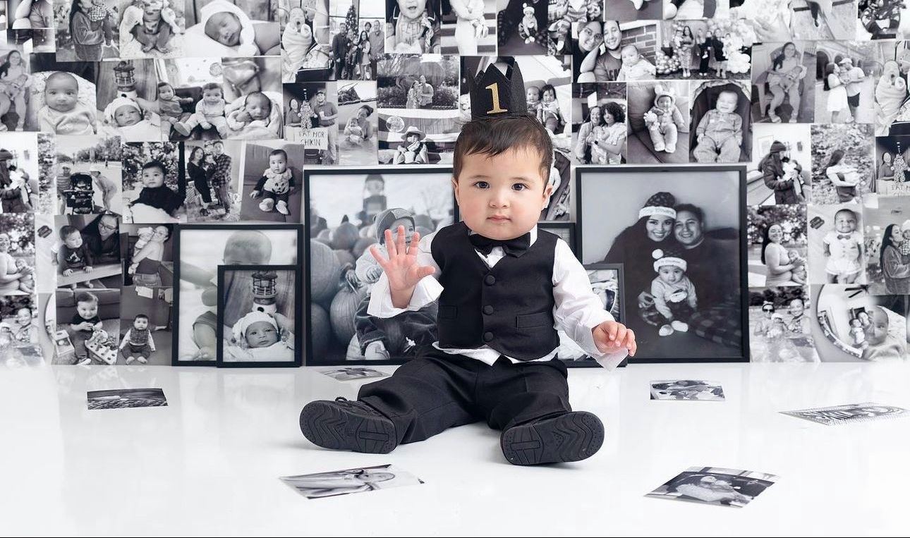 A baby in formal attire sits on white floor surrounded by black and white photographs on the wall behind them.