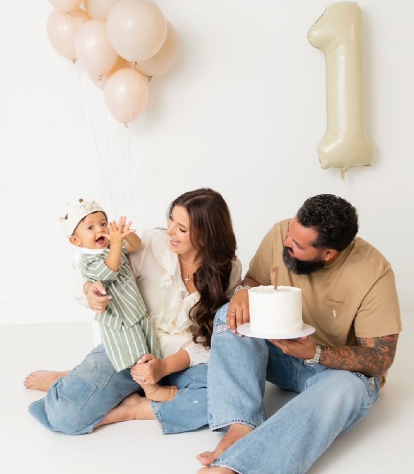 Family celebrating first birthday with cake and peach balloons in minimalist white studio setting.