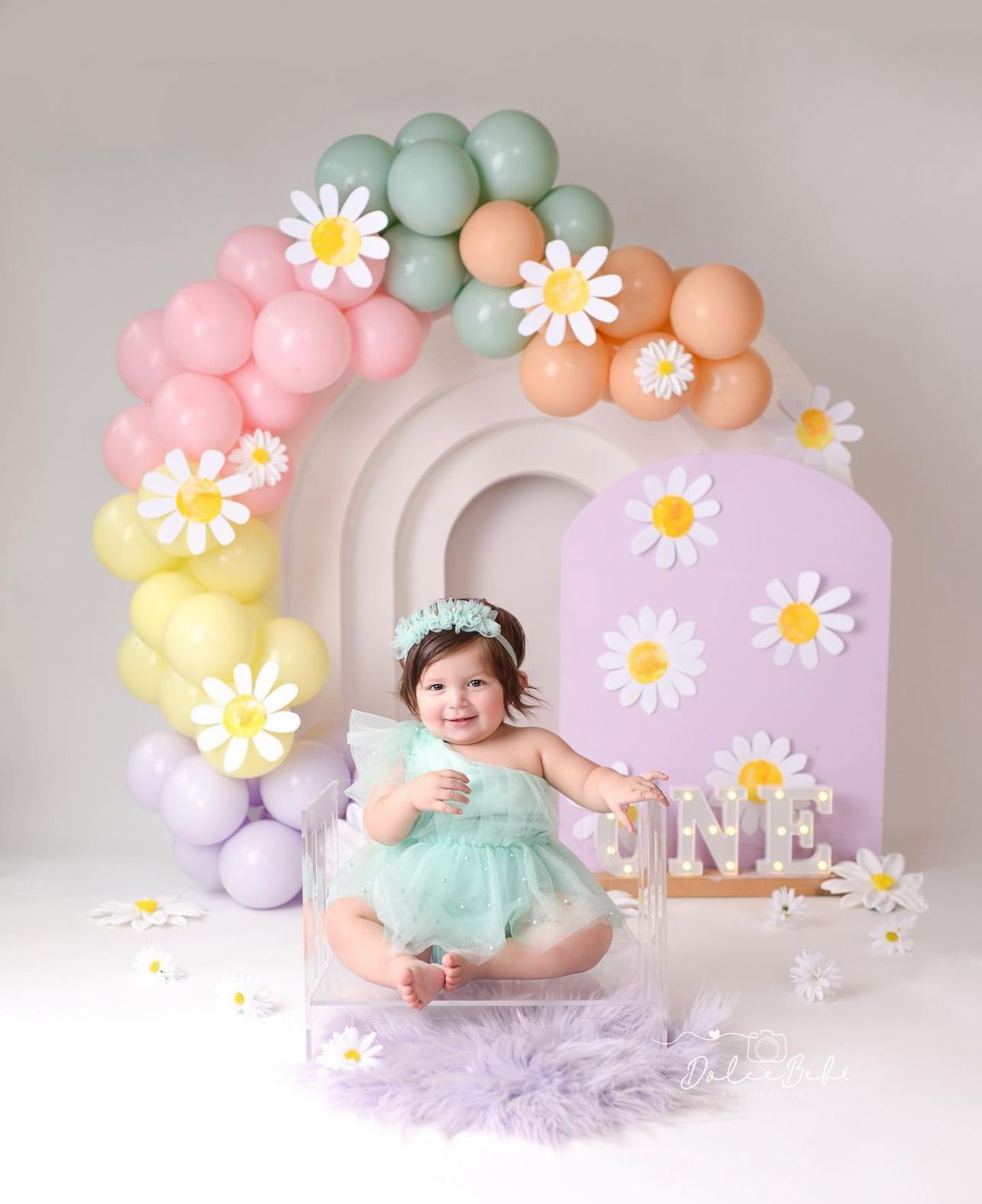 Pastel balloon arch with daisies decorates a dreamy first birthday setup featuring soft purple and mint colors.