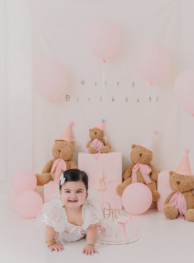 Sweet first birthday setup with teddy bears, pink balloons and decorations against white backdrop.