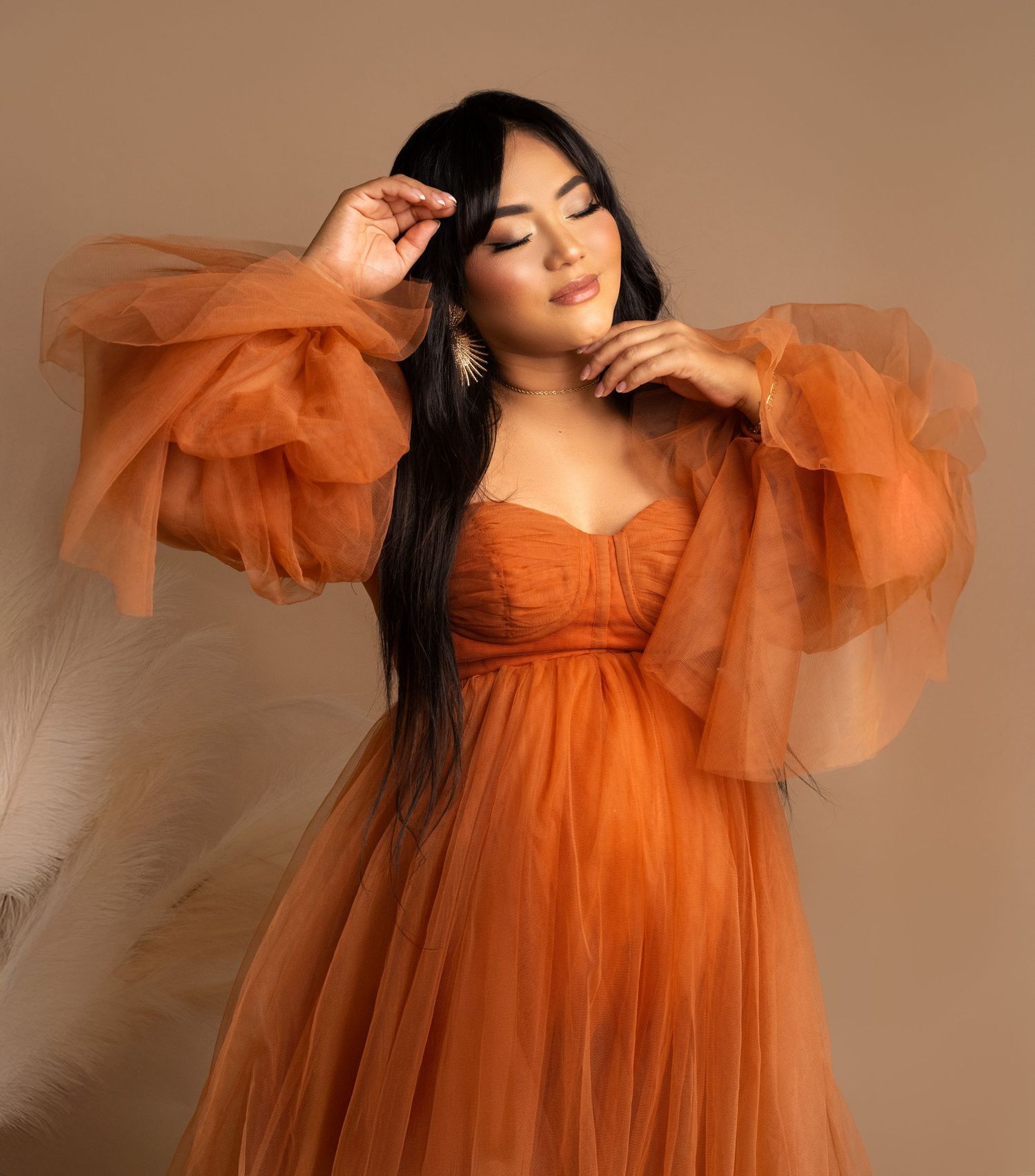 A woman poses in a flowy orange tulle gown with dramatic puff sleeves against a neutral studio backdrop.