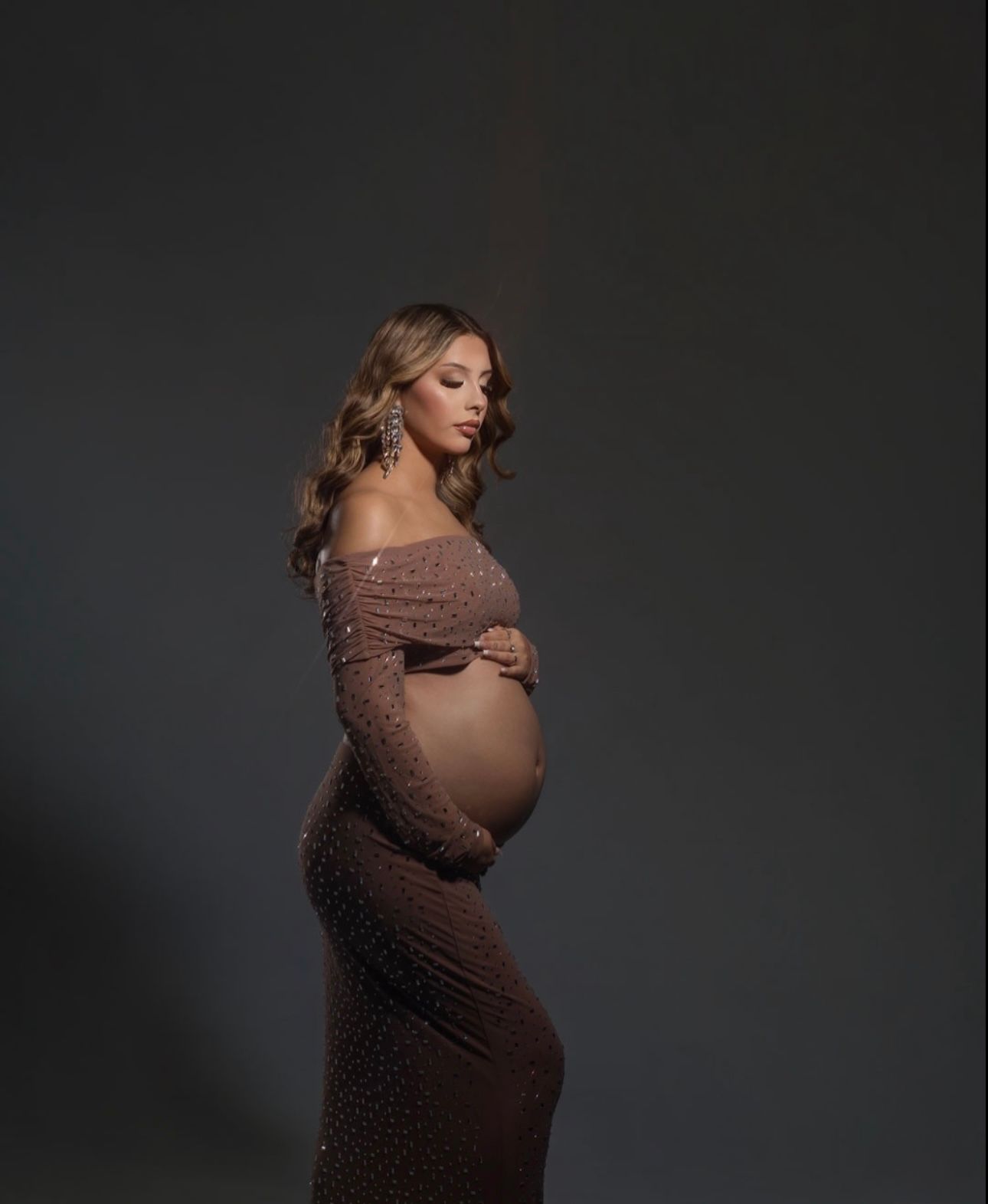 Pregnant woman in a fitted sparkly mauve off-shoulder maternity dress posing in a professional studio.