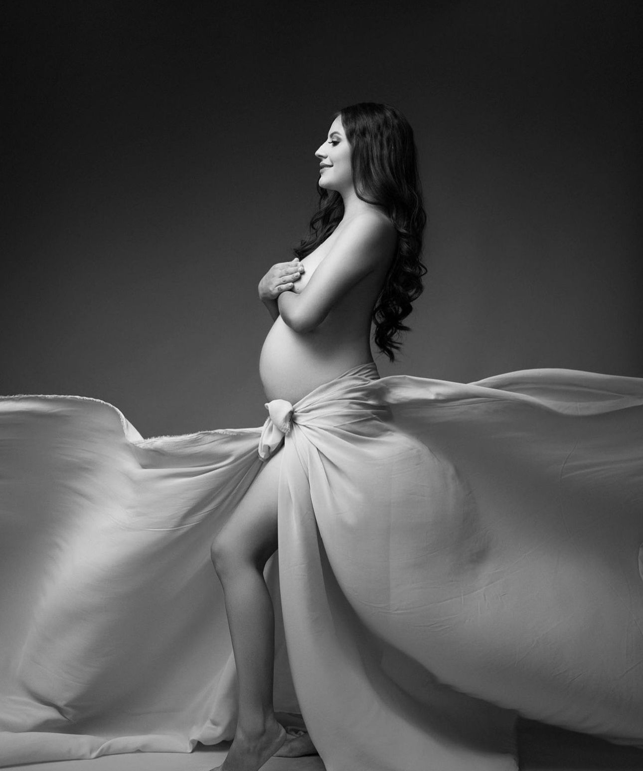 Black and white maternity photo of a pregnant woman draped in flowing fabric, posing elegantly in a studio setting.