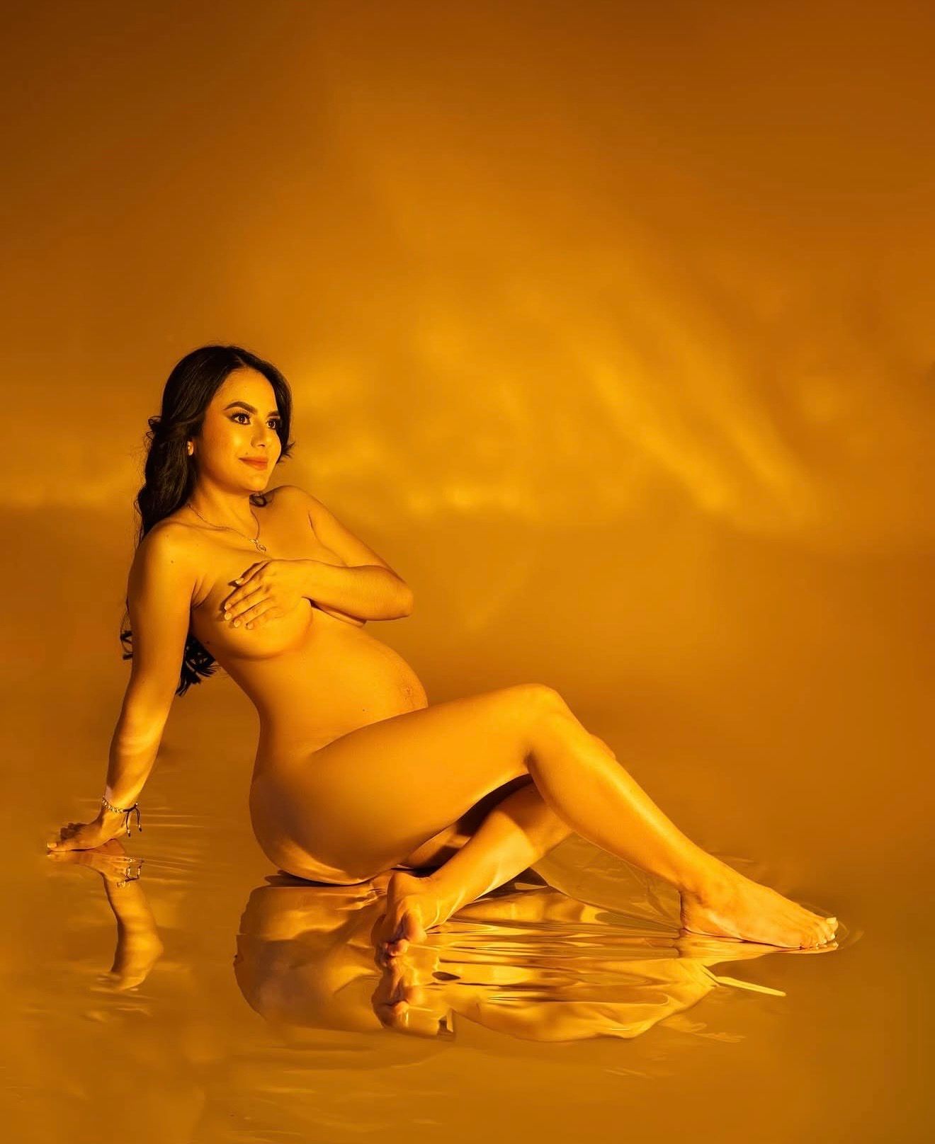 Pregnant woman posing gracefully in golden amber light, seated with reflective water surface below.
