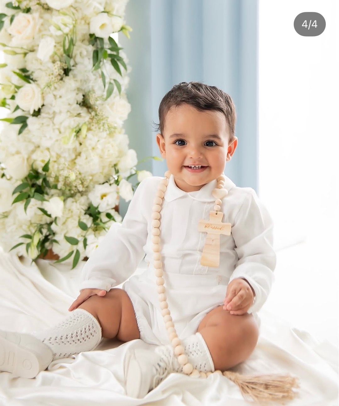 Smiling baby boy in white outfit holding wooden beaded rosary, seated among white floral arrangements.