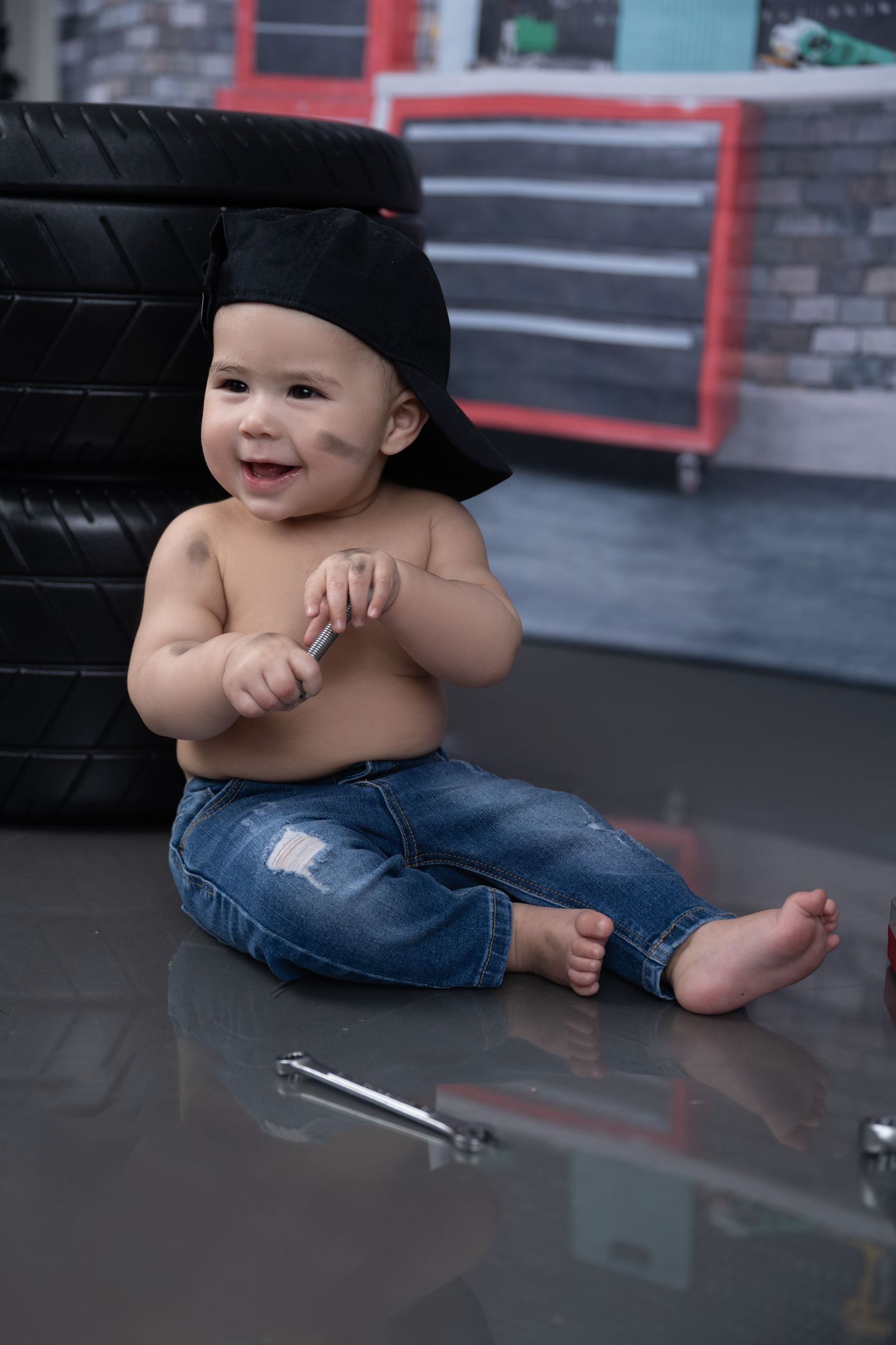 A happy toddler in jeans sits barefoot next to stacked tires in an auto repair shop setting.