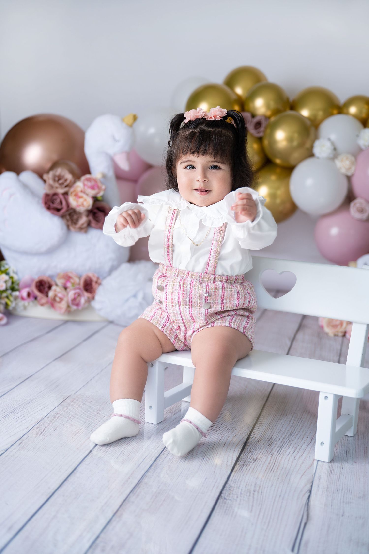 Baby in pink romper sits on white bench with balloon and floral backdrop.