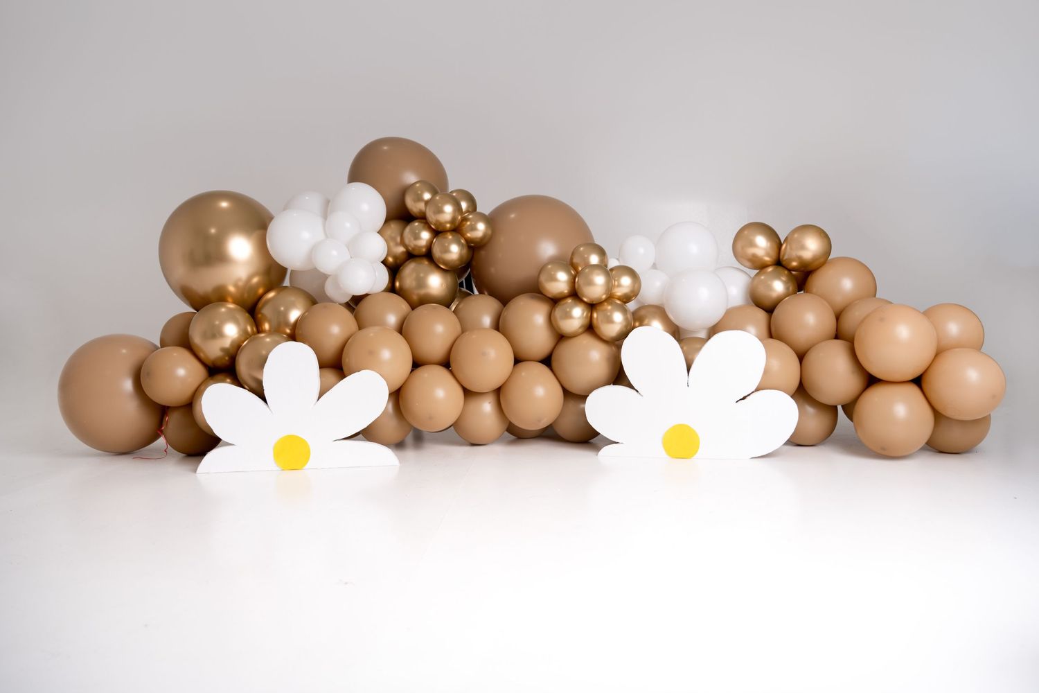 Decorative balloon arch arrangement in neutral tones with white daisy flowers against white background.
