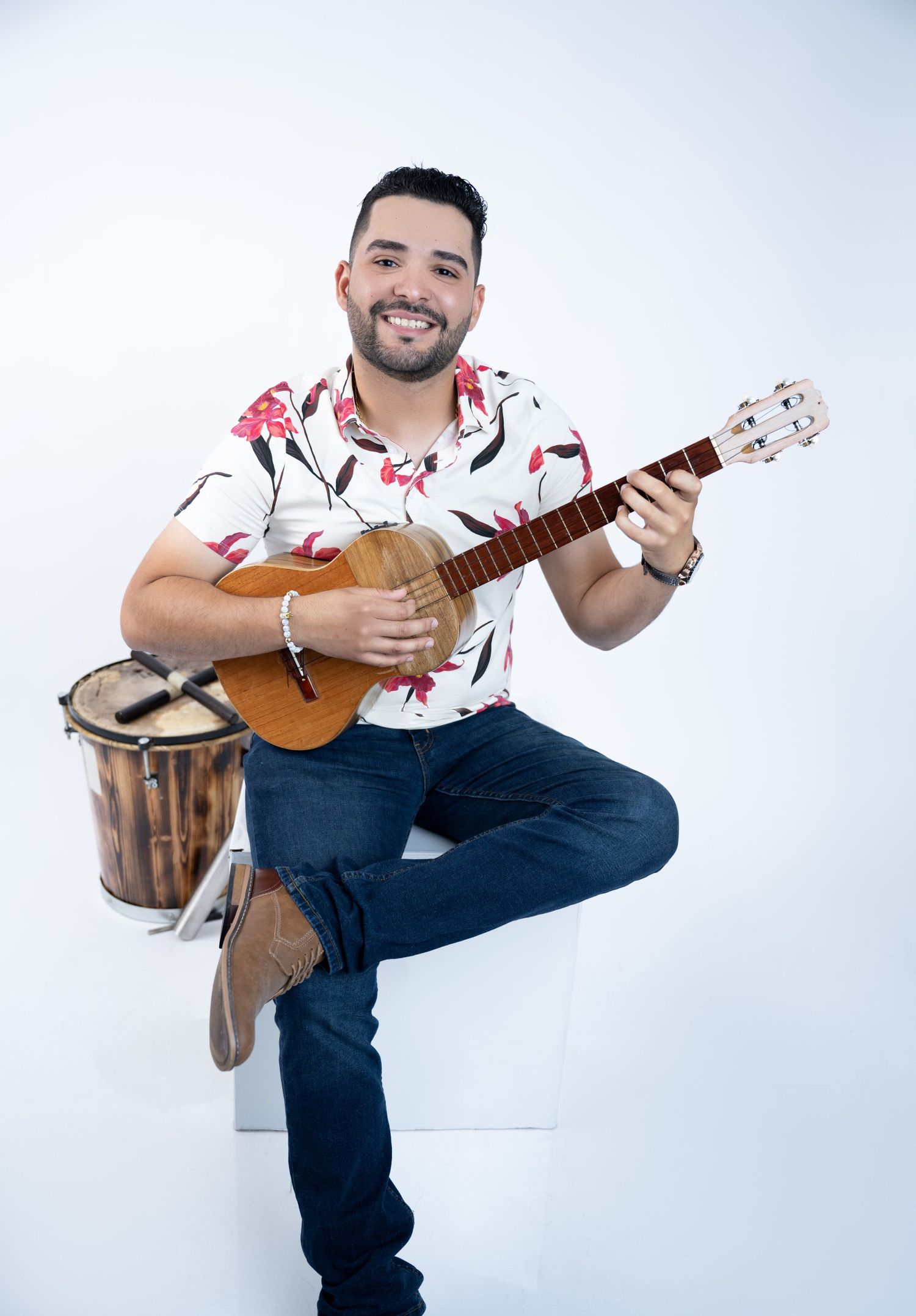 Musician in floral shirt playing ukulele while seated on white cube.