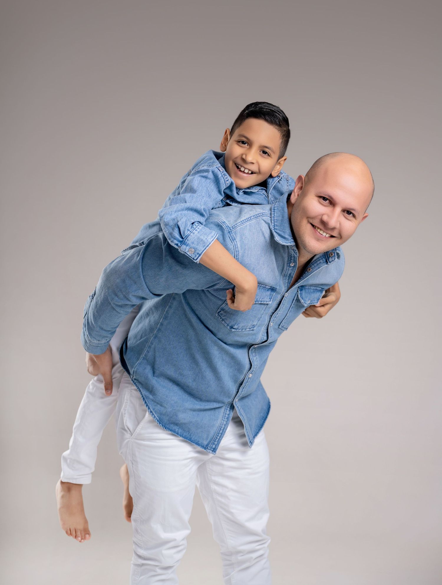 A bald man in a blue denim shirt gives a smiling boy a piggyback ride, both wearing white pants.