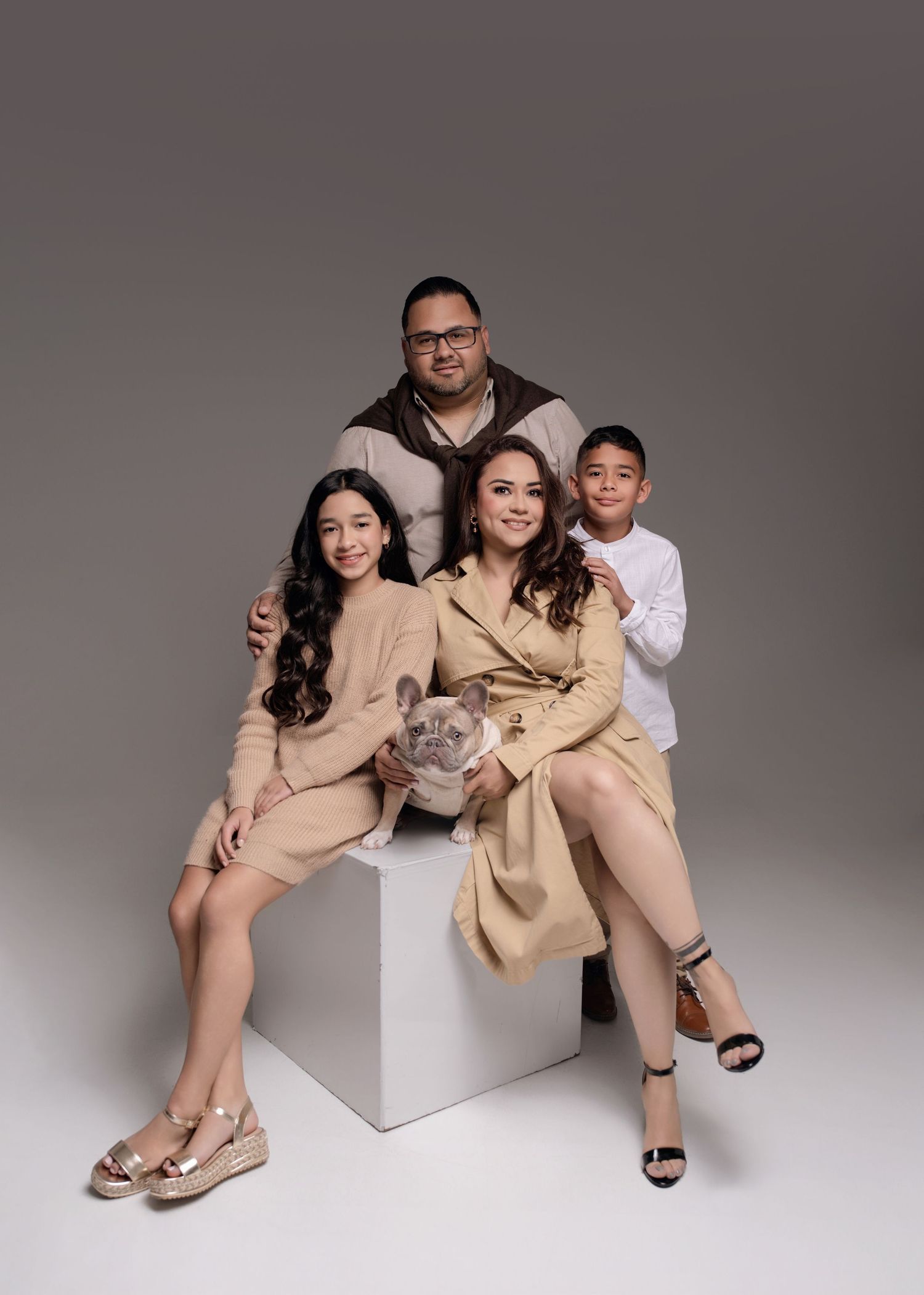 Family of four in beige outfits posing with a French Bulldog puppy in a professional studio portrait.