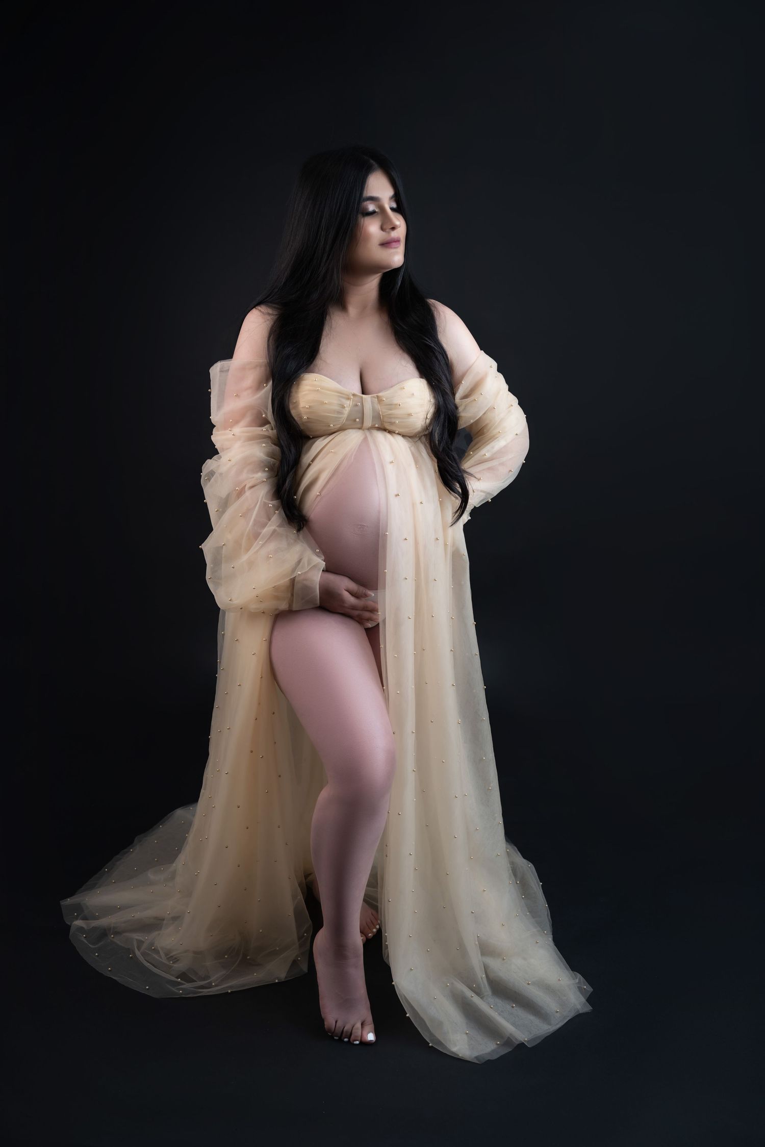 Pregnant woman in sheer cream robe and lingerie posing gracefully against dark background in maternity photo session.