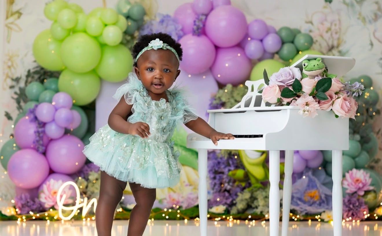 Toddler girl in mint dress stands by white toy piano with colorful balloon arch backdrop at birthday photoshoot.