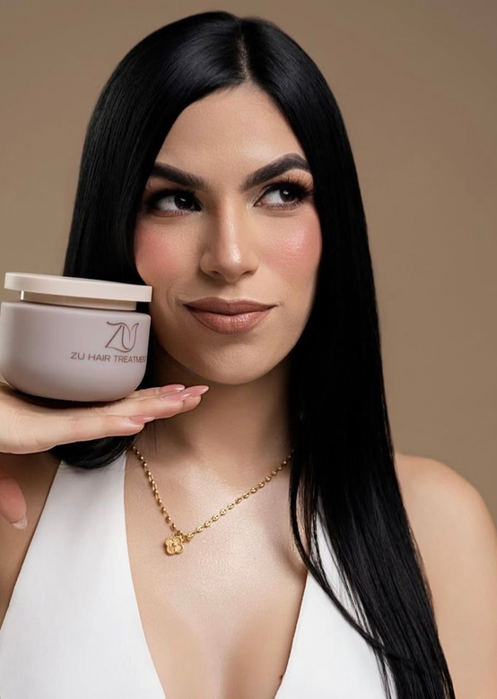 Woman with long straight black hair holding a jar of ZU Hair Treatment against a tan background, wearing a gold necklace and white top.