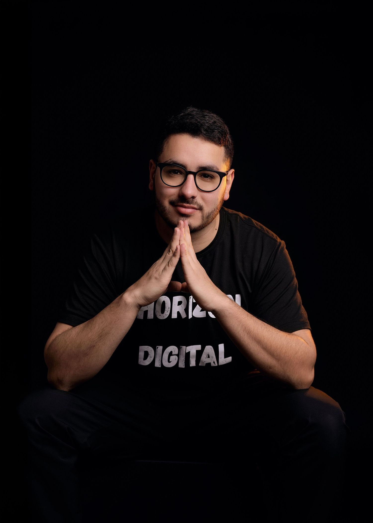 Man with glasses and a beard wearing a black Horizon Digital t-shirt, sitting with hands pressed together against a dark background.