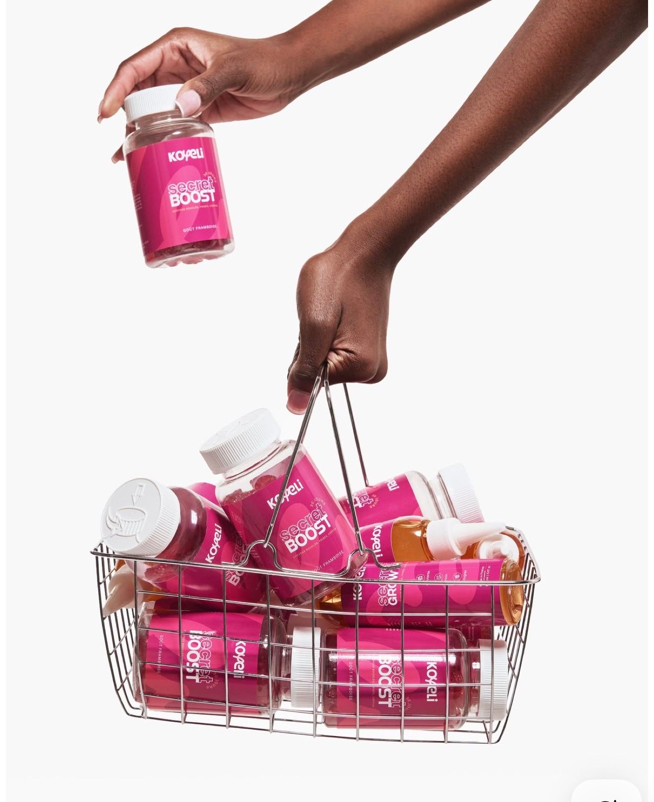 Hand placing a Kopari Beauty Boost supplement bottle into a wire basket filled with pink Kopari beauty products.