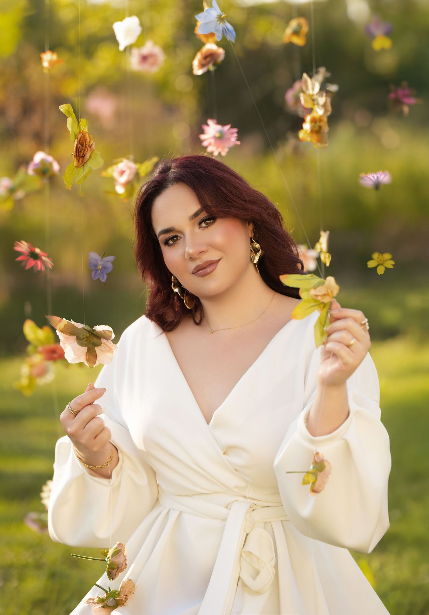 Woman in white wrap dress holding flowers outdoors with colorful floral decorations hanging in background.