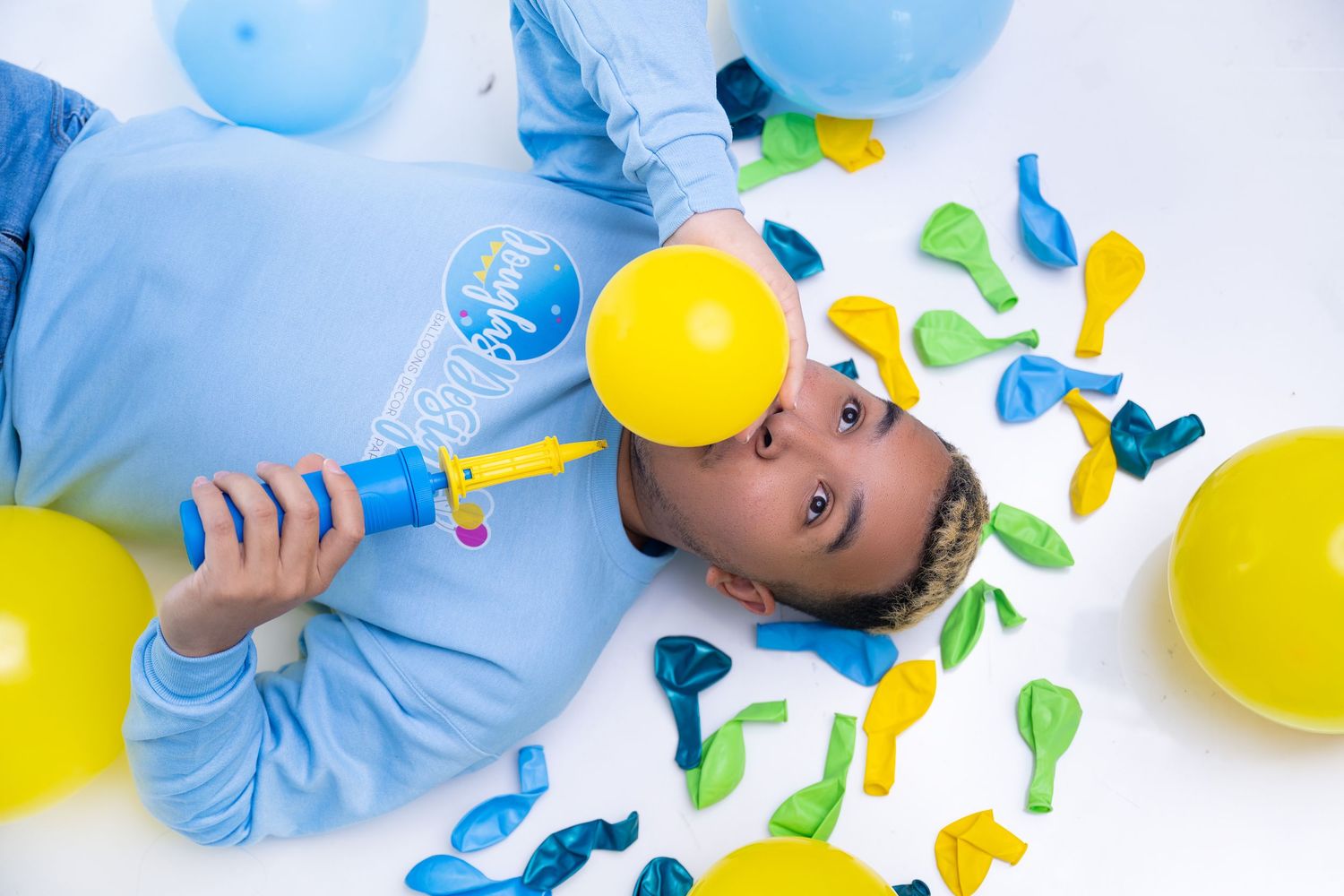 Child in blue hoodie lying on floor surrounded by colorful yellow, green, and blue balloons holding a balloon pump.
