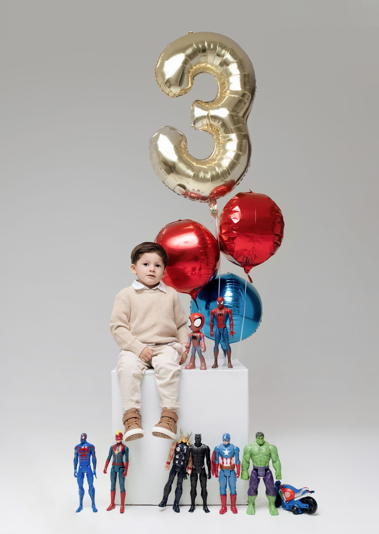 Boy sitting on white cube with Marvel superhero toys and balloons celebrating 3rd birthday.