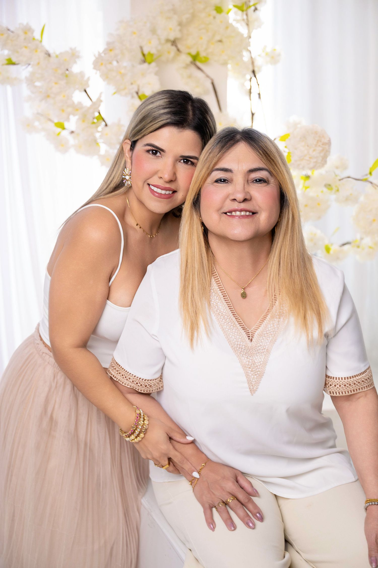 Two smiling women in white and blush outfits pose together in front of white floral decorations.