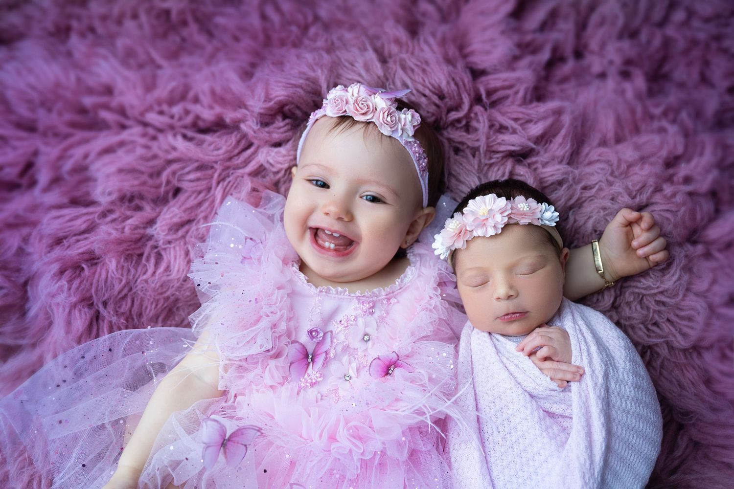 Two baby girls in pink outfits and floral headbands pose on a purple fur rug, one smiling and one sleeping newborn.
