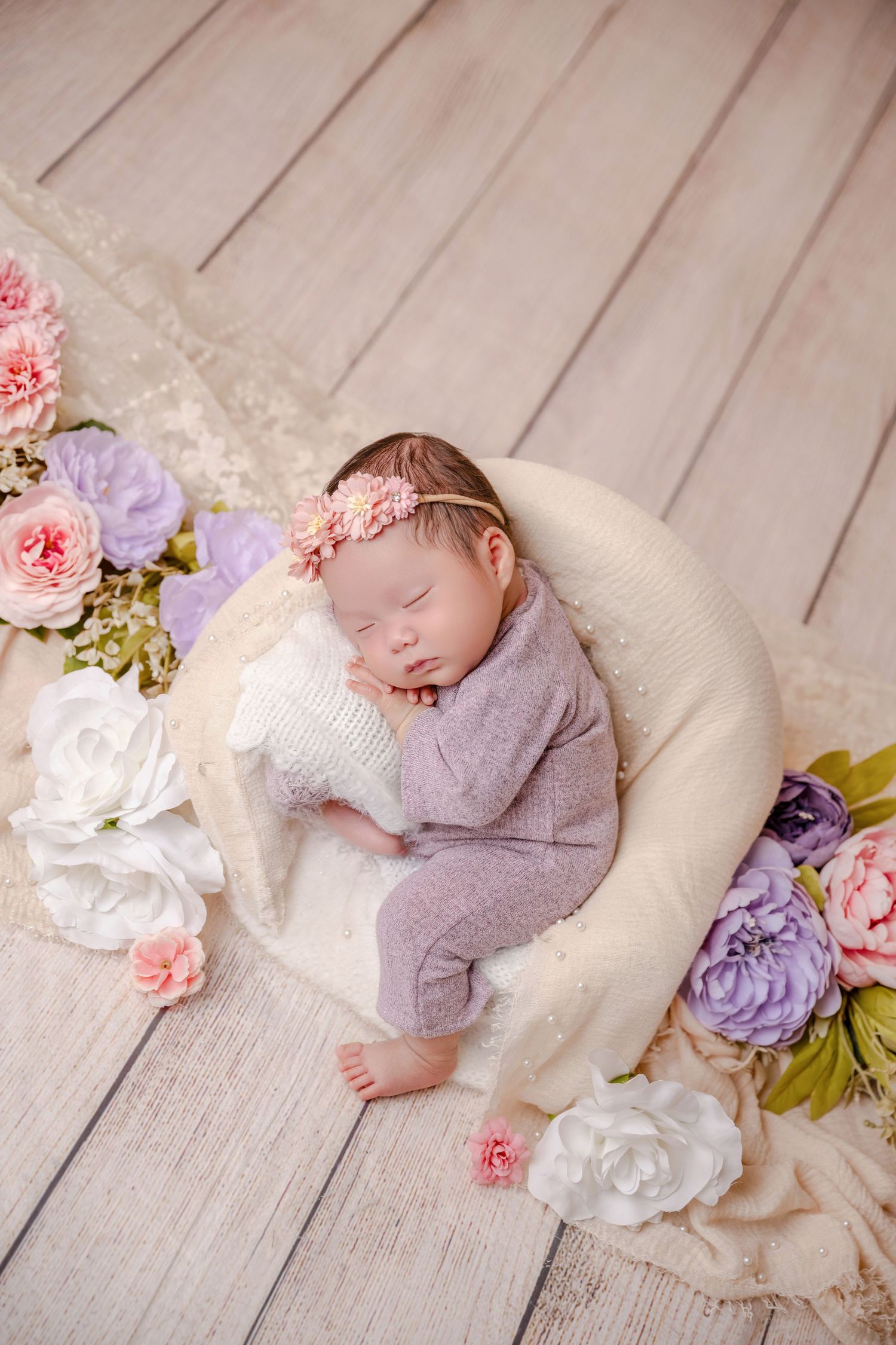 Sleeping newborn baby girl in lavender outfit with floral headband resting in cream bowl surrounded by pink and purple flowers.
