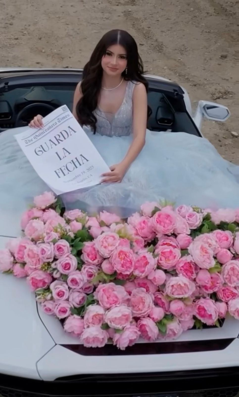 Young woman in blue gown holding Save the Date sign, posing with pink roses on white convertible car.
