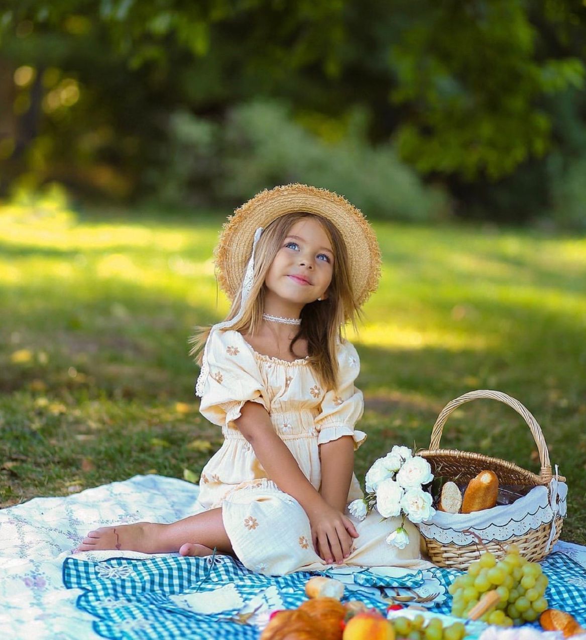 Child in white dress and straw hat having a picnic on blue gingham blanket with pumpkins and flowers outdoors.