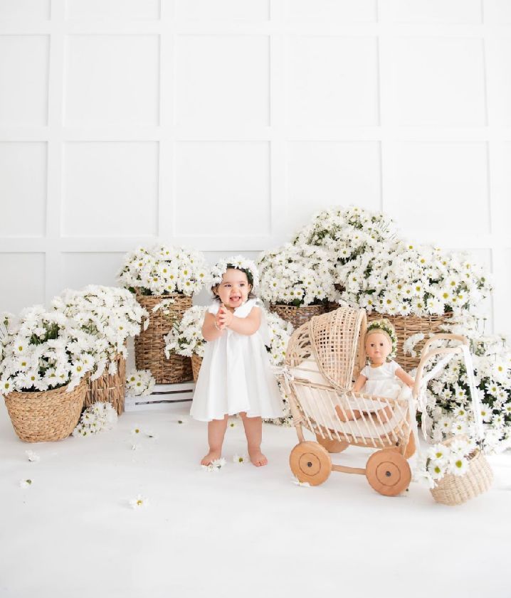 A baby sits in a vintage wicker pram surrounded by white flowers and baskets in an all-white studio setting.