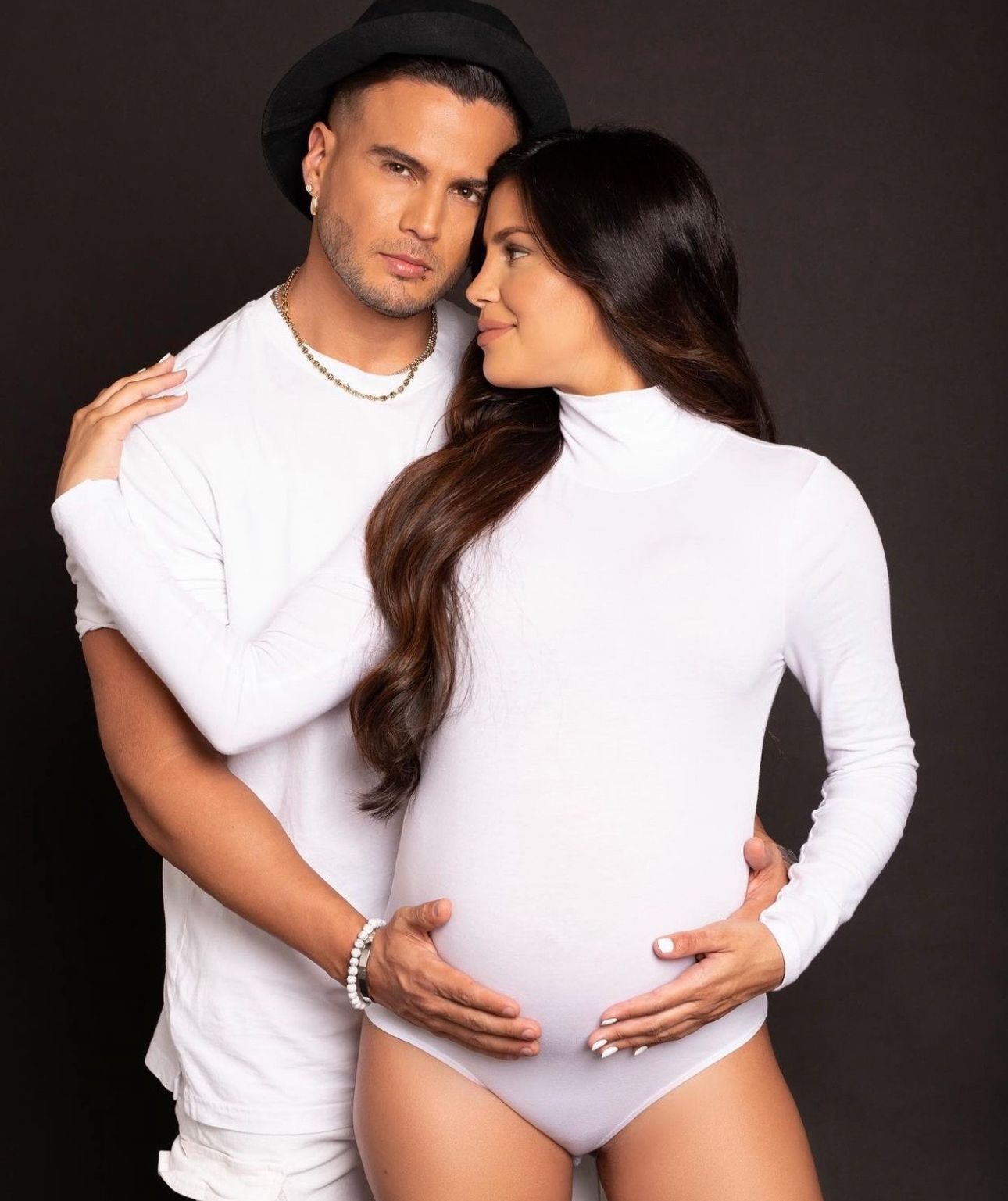 Couple in white outfits posing for maternity photo shoot, woman cradling baby bump, man in black hat embracing her.