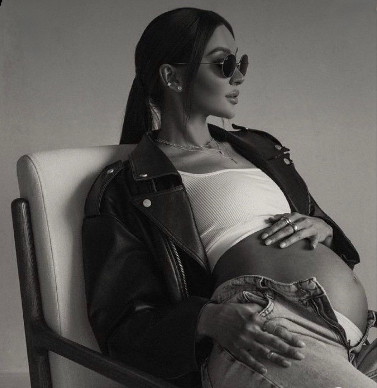 Black and white photo of a pregnant woman in sunglasses, leather jacket, and crop top sitting in a white chair.