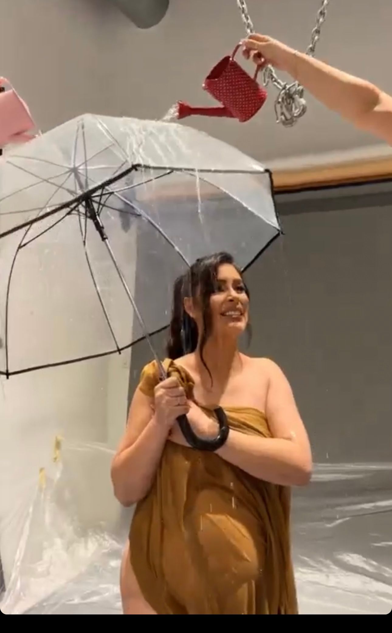 Pregnant woman in gold dress holds clear umbrella indoors during a creative maternity photoshoot with decorative watering cans.