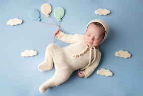Sleeping baby in cream knit outfit lying on blue backdrop with paper cloud decorations and pastel balloons.