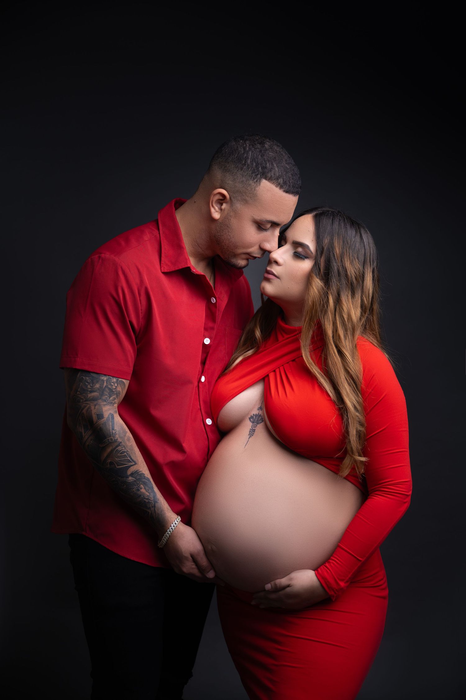 A romantic maternity photoshoot featuring a couple in red outfits against a dark background.