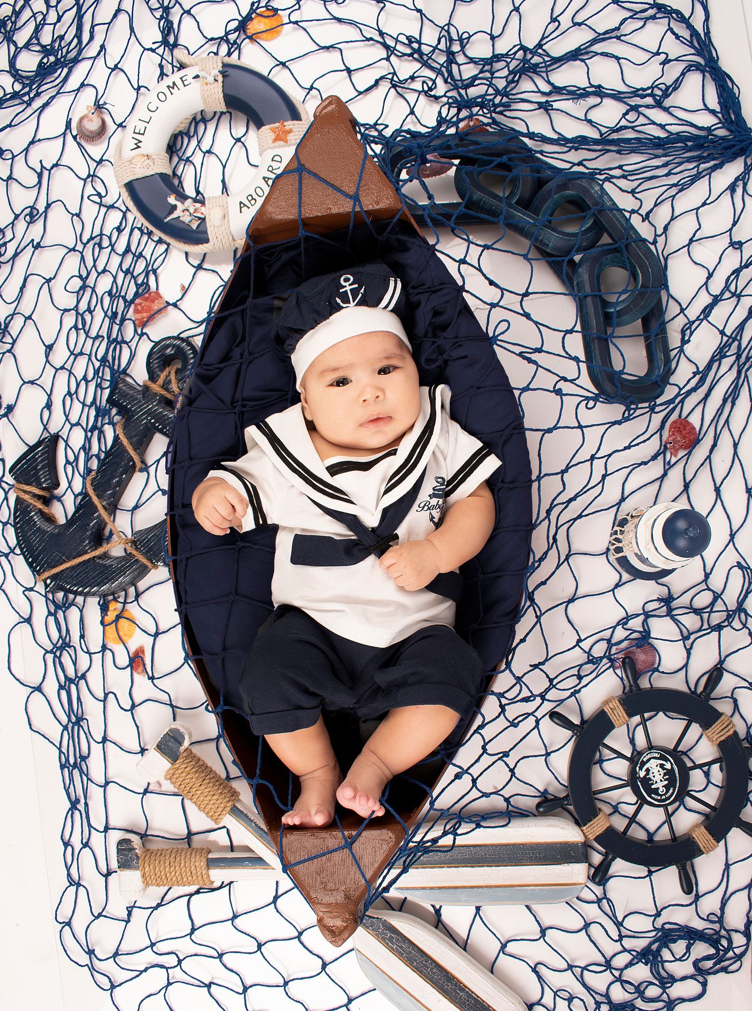 Baby in nautical outfit lying on blue fishing net with maritime decorations.